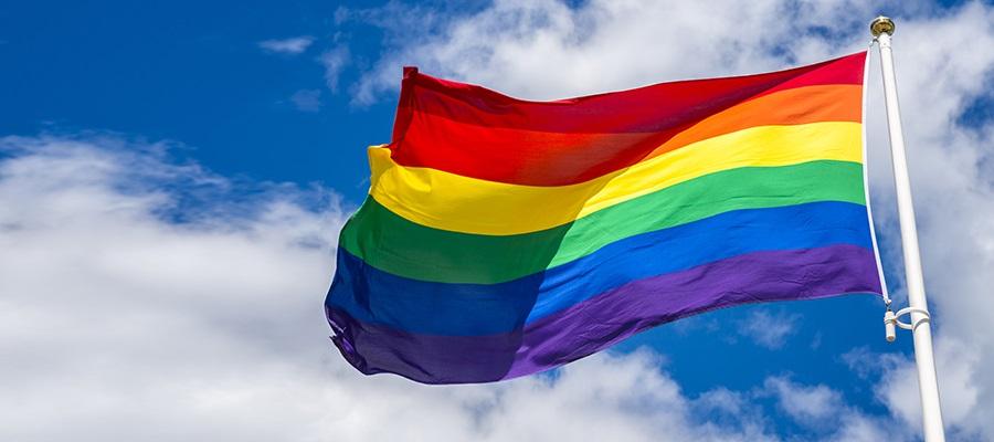 Gay Pride flag flying against a blue sky with white clouds.